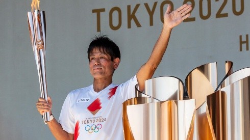 Um dos carregadores da tocha olímpica posa empunhando-a e acenando durante evento em Tóquio (Foto: Getty Images)